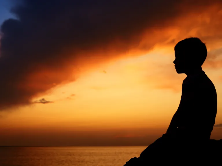 A young boy's seated profile is silhouetted against a dramatic orange sunset