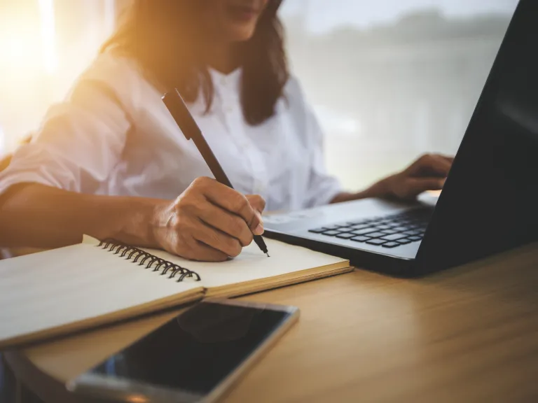 A woman with mid-length dark hair sitting in front of a laptop while writing in a notebook